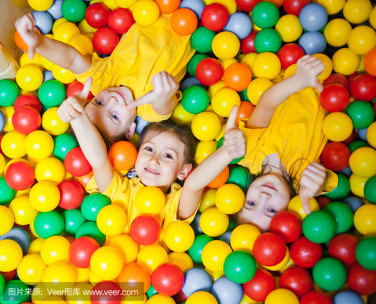 三个快乐的小女孩在球坑快乐地微笑着对着镜头和显示拇指,而在儿童游戏中心玩得很开心。俯视图